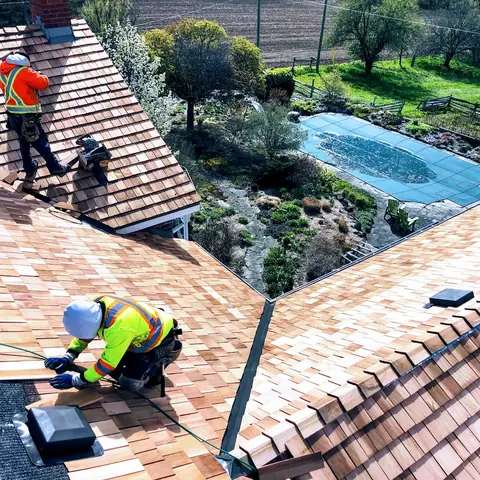 Two roofers in high-vis laying new cedar shakes course by course on a country home
