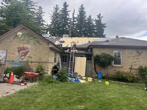 Testarossa Roofing crew re-roofing a bungalow with a hand-painted mural on the gable, ladder up and new roof sheathing going down