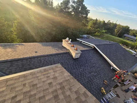 Finished charcoal shingle roof with a stone chimney freshly flashed and capped, golden hour light through the trees