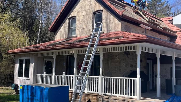 Roofer installing a red metal tile roof on a stone farmhouse, secured on a safety line