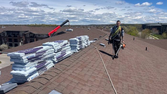 Roofer harnessed and walking a steep roof ridge with bundled shingles staged nearby, aerial view